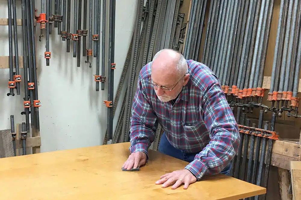 Bob sanding a wooden tabletop by hand in his woodshop, surrounded by pipe clamps hanging on the wall.